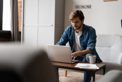 Young male adult sitting on his couch and looking at his laptop